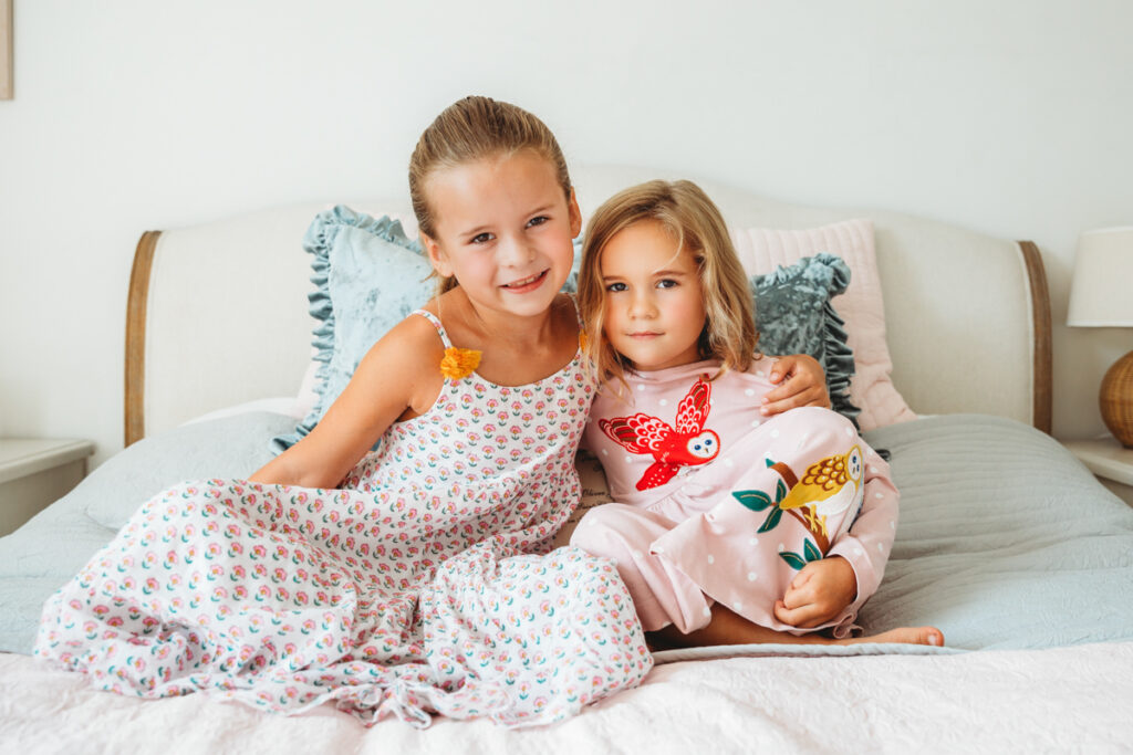 Two little sisters sitting on bed hugging each other for a photo.