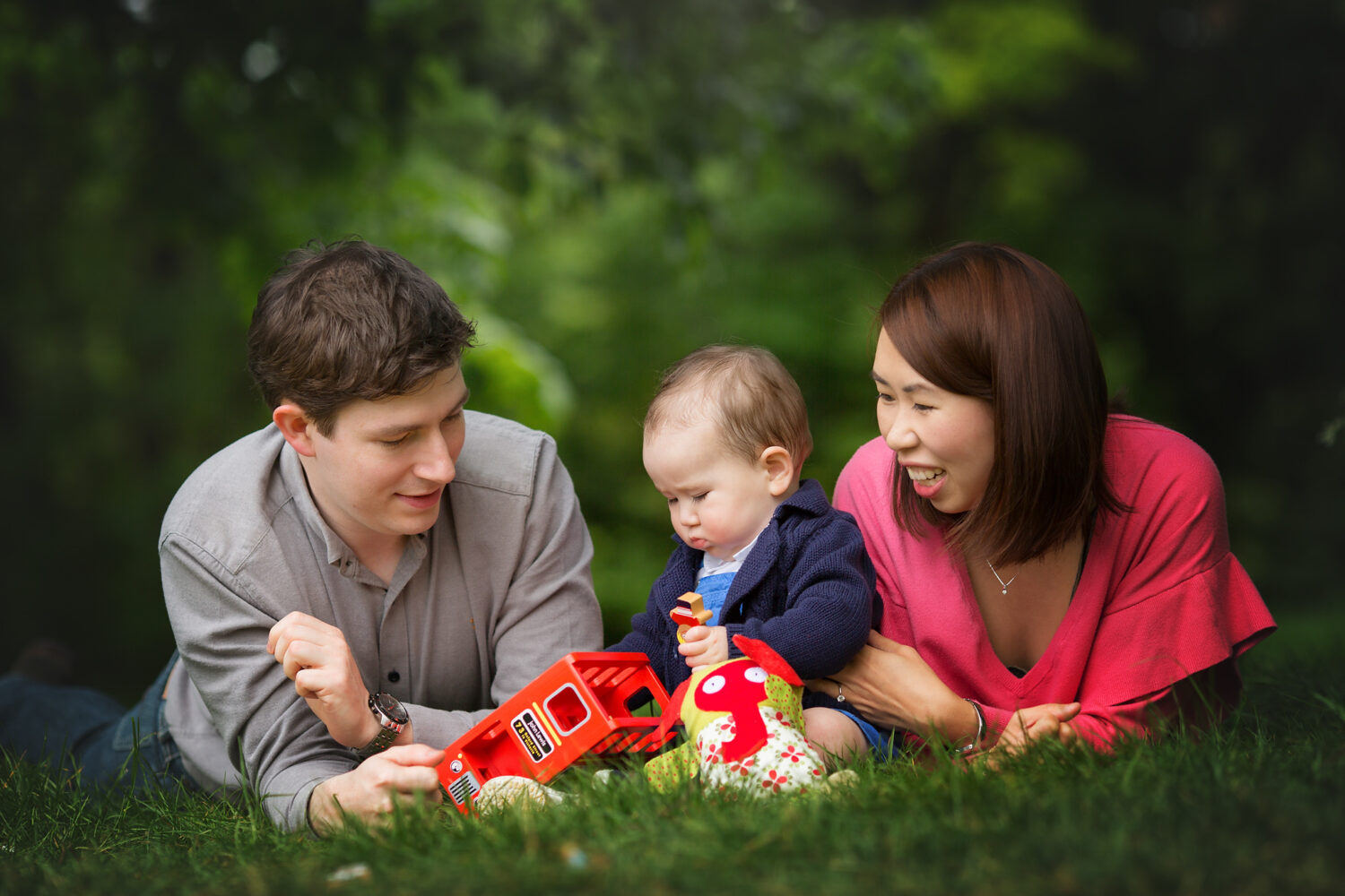 Mum dad and little baby lying on the grass playing with toys.