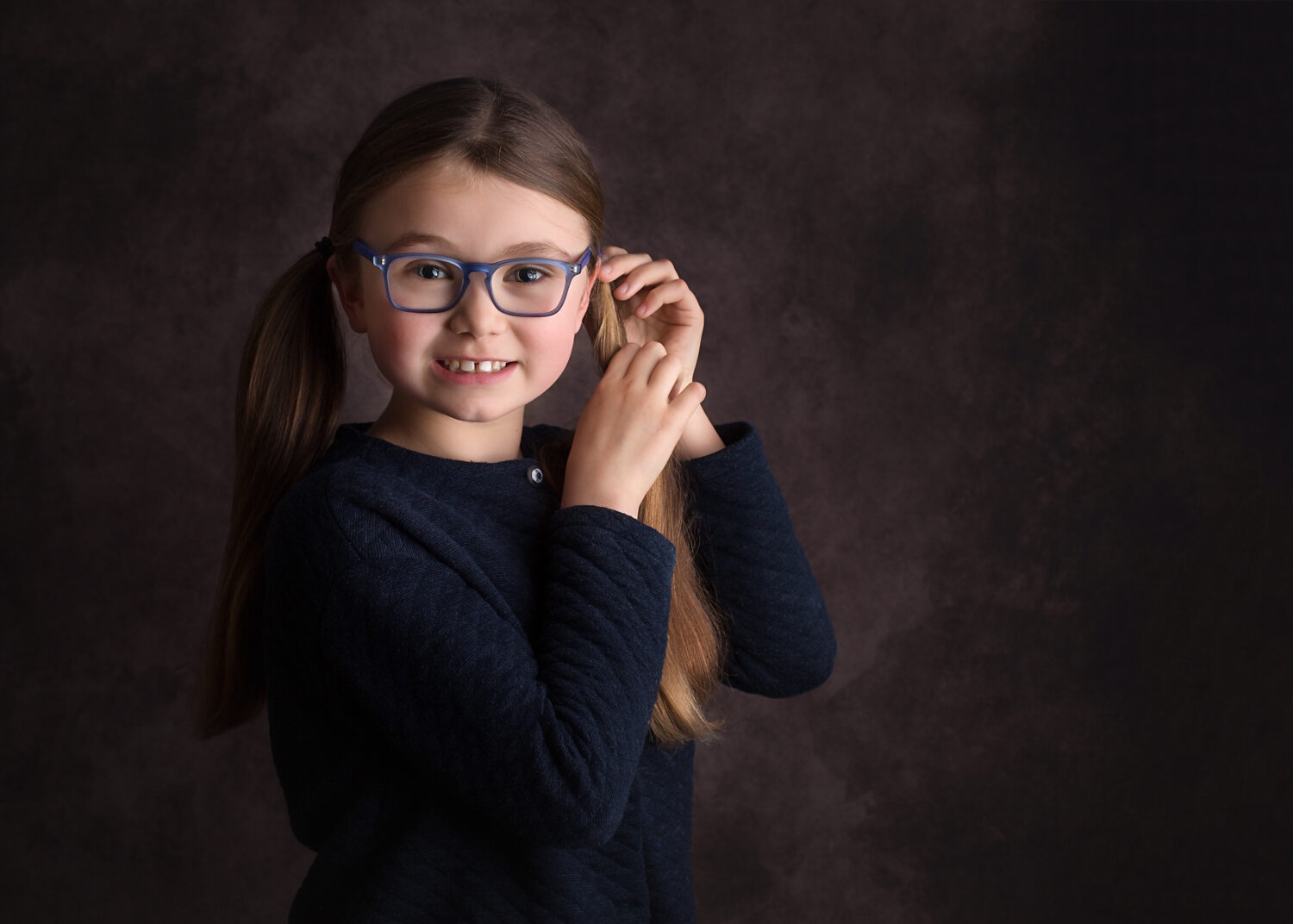 Girl touching her, in a brown photography backdrop, looking and smiling at the camera in a blue sweater. She's wearing glasses and she has 2 pretty pony tails.