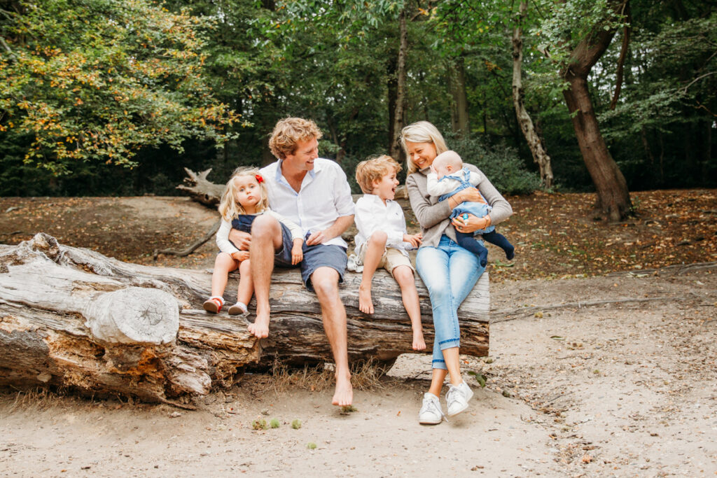 Mum, dad, a little boy, and girl sitting on a log in Wimbledon park laughing and having fun. Mum is also holding their baby.