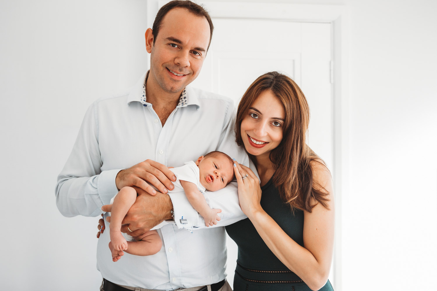 Mum and dad next to each other holding their newborn baby and smiling at the camera.