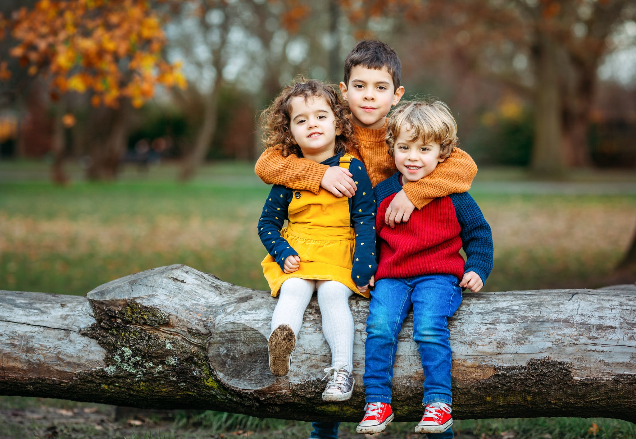 3 kids sitting on a log and hugging each other at Wandsworth Common.